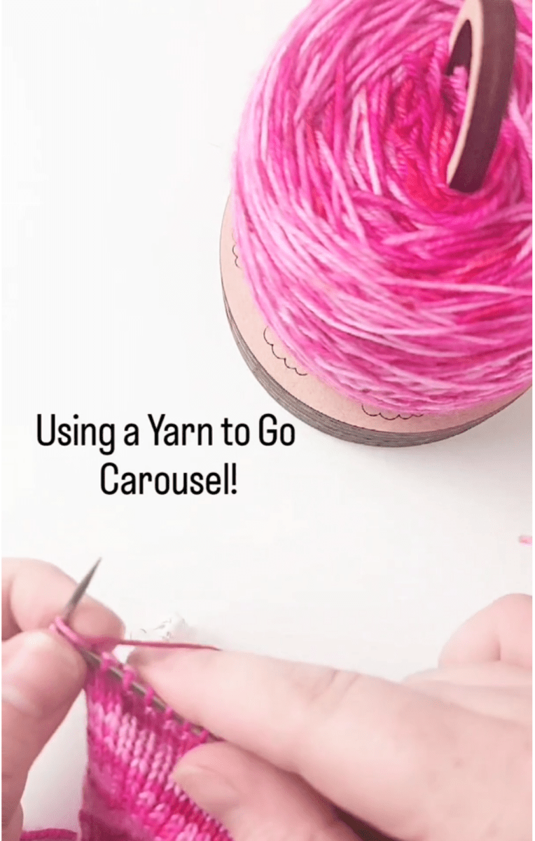 Close-up of hands knitting with pink yarn. A yarn cake rests on the Twice Sheared Sheep Yarn To Go Carousel, Portable Yarn Butler. Text reads: Tangle-free knitting with the Yarn To Go Carousel!.