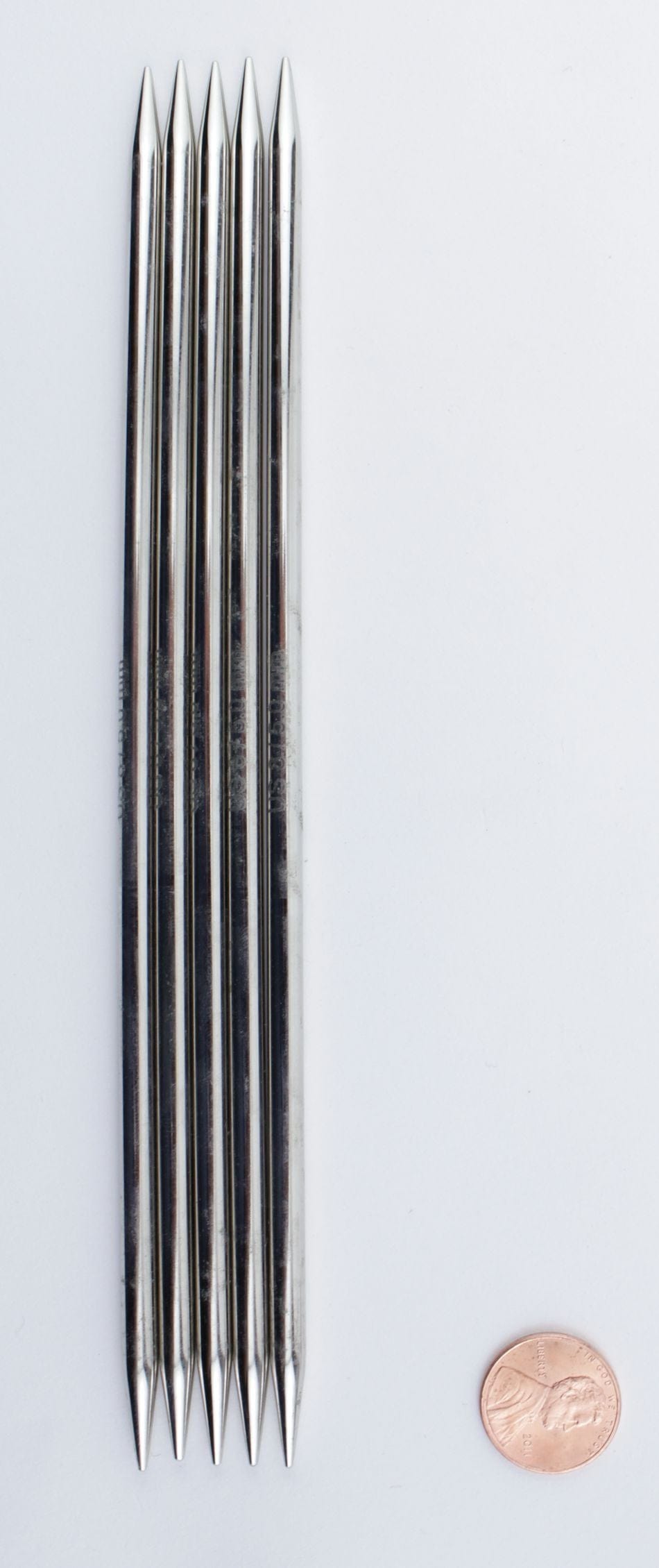 Six Accessories Unlimited Knit Pro Nova Metal Double-Point Knitting Needles, each 6 inches long, are arranged in a row on a white surface with a penny beside them for size comparison.