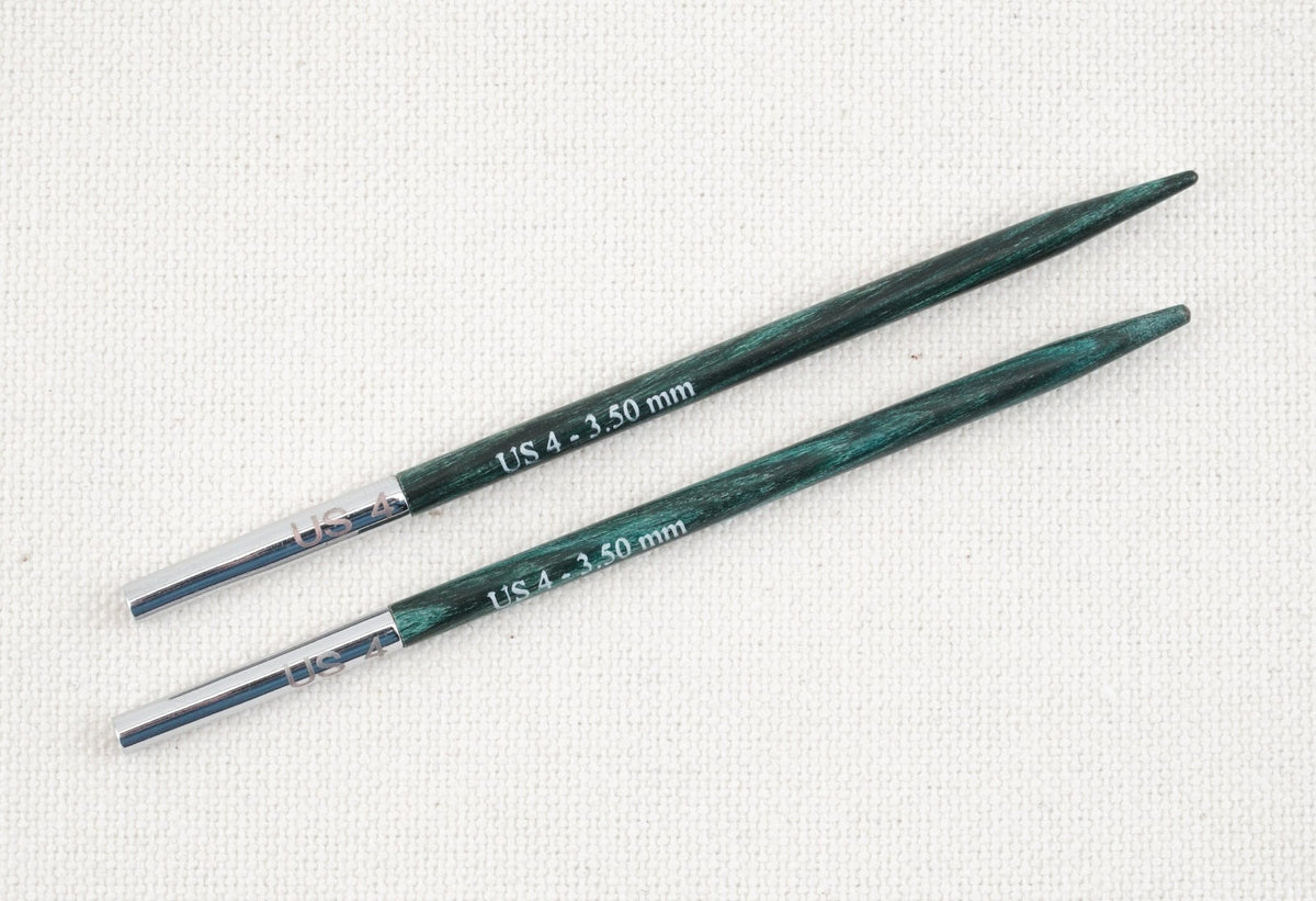 Two dark green Knit Pro (Knitters Pride) Dreamz Interchangeable Knitting Needle Tips by Accessories Unlimited, size US 4 (3.50 mm), lay parallel on a textured cream fabric background, with silver metal bases visible.