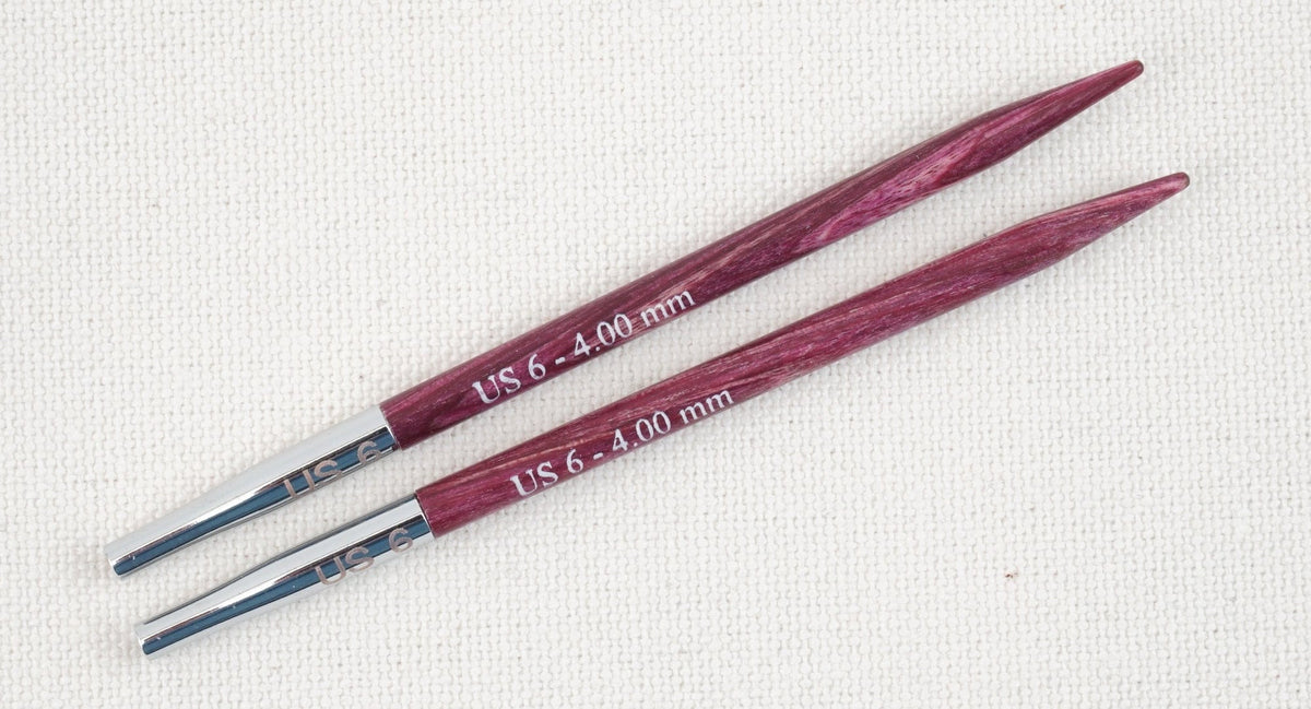A pair of Accessories Unlimited Knit Pro Dreamz interchangeable knitting needle tips, US 6 (4.00 mm), with purple laminated birch wood and silver metal ends, are shown parallel on a white fabric background.