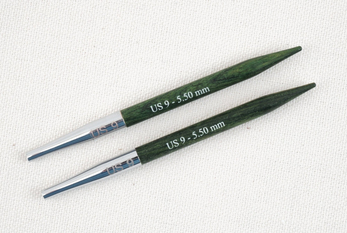 Two green and silver Knit Pro (Knitters Pride) Dreamz Interchangeable Knitting Needle Tips by Accessories Unlimited, size US 9 – 5.50 mm, lie parallel on a white textured surface. Crafted from smooth laminated birch for comfortable stitching.