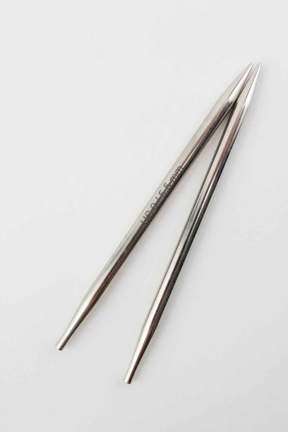 Two metal Knit Pro (Knitters Pride) Nova 5" interchangeable knitting needles by Accessories Unlimited rest diagonally on a white surface, their pointed, reflective tips visible and one needle showing engraved text near the top.