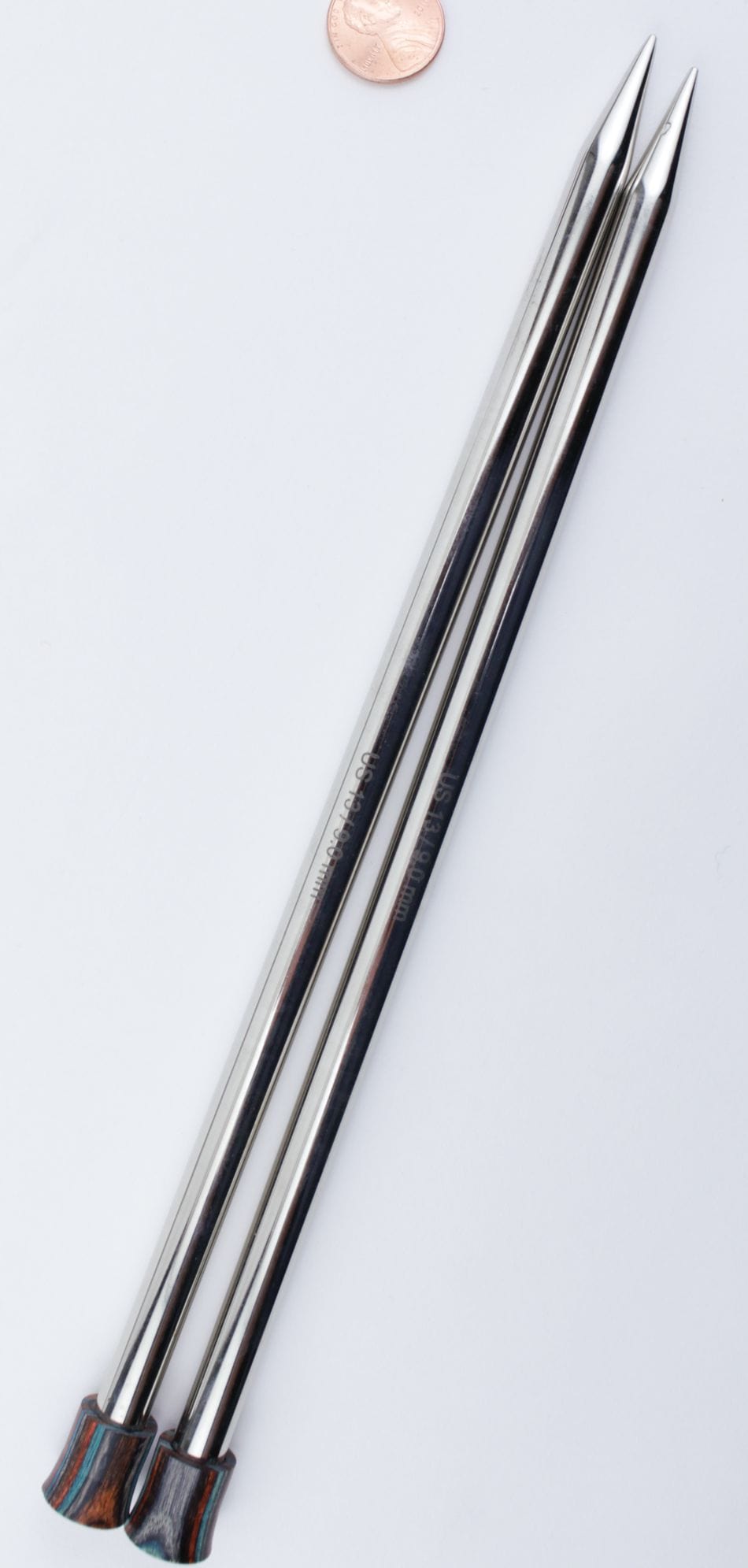 Two Knit Pro (Knitters Pride) Nova Metal Single Point Knitting Needles (10" or 14") by Accessories Unlimited are placed parallel on a white surface, with a penny above them for size comparison.