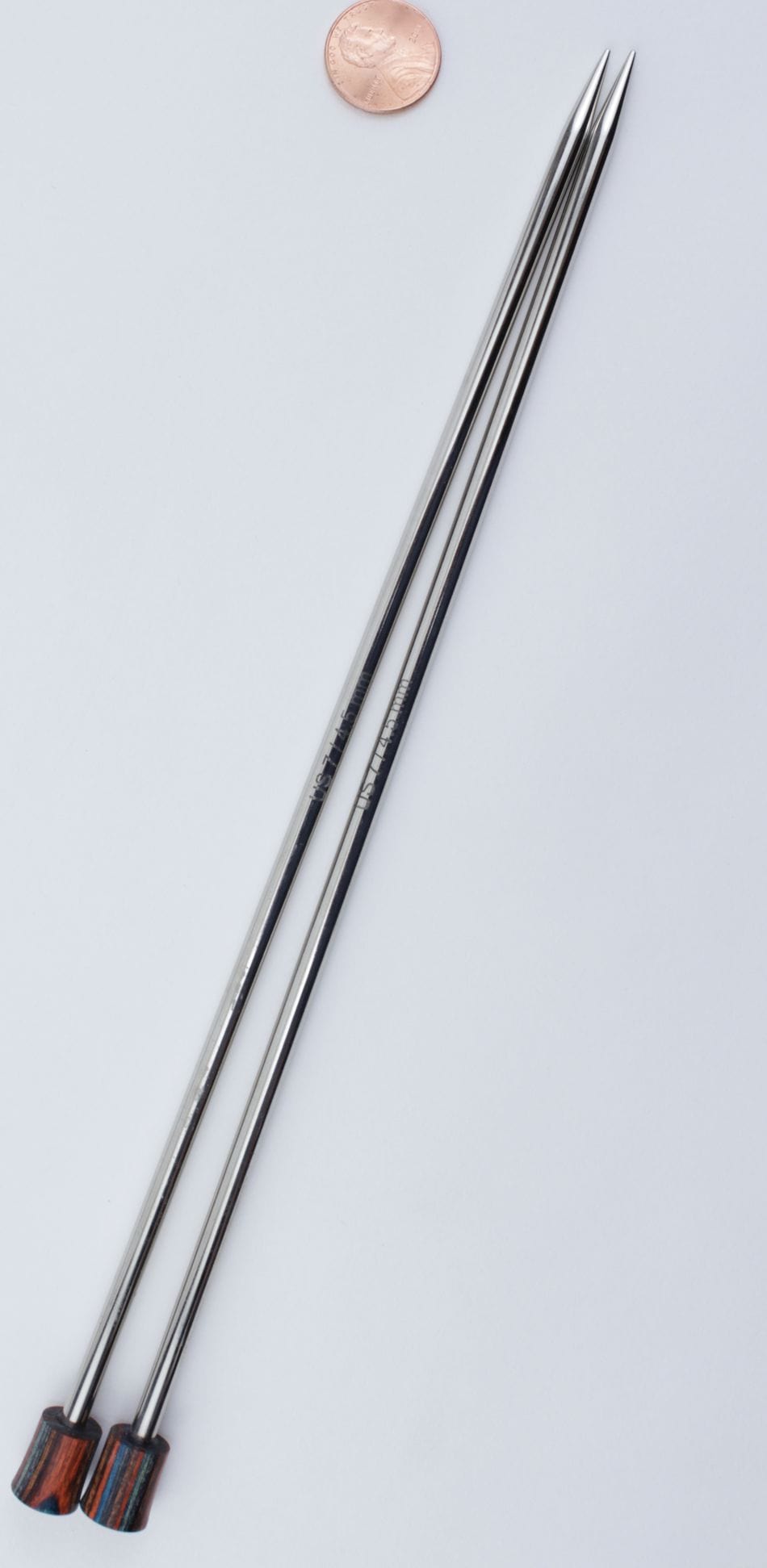 Two silver Knit Pro Nova Metal Single Point Knitting Needles (10" or 14") by Accessories Unlimited, with pointed tips and wooden handles, are placed parallel on a white surface, with a penny above them for scale.