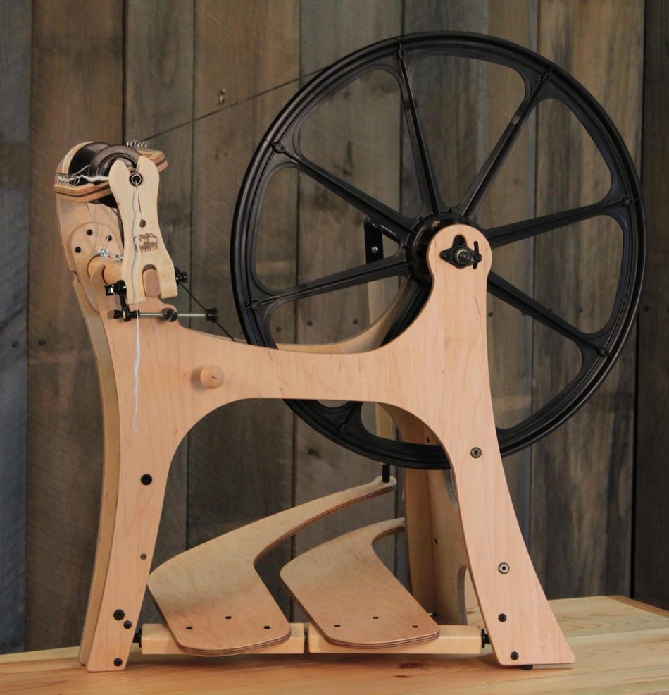 A Flatiron Spinning Wheel, Double-Treadle by Schacht Spindle Co., featuring a large black drive wheel, is displayed on a wooden surface in front of rustic wood paneling. Temporarily out of stock with Schacht.